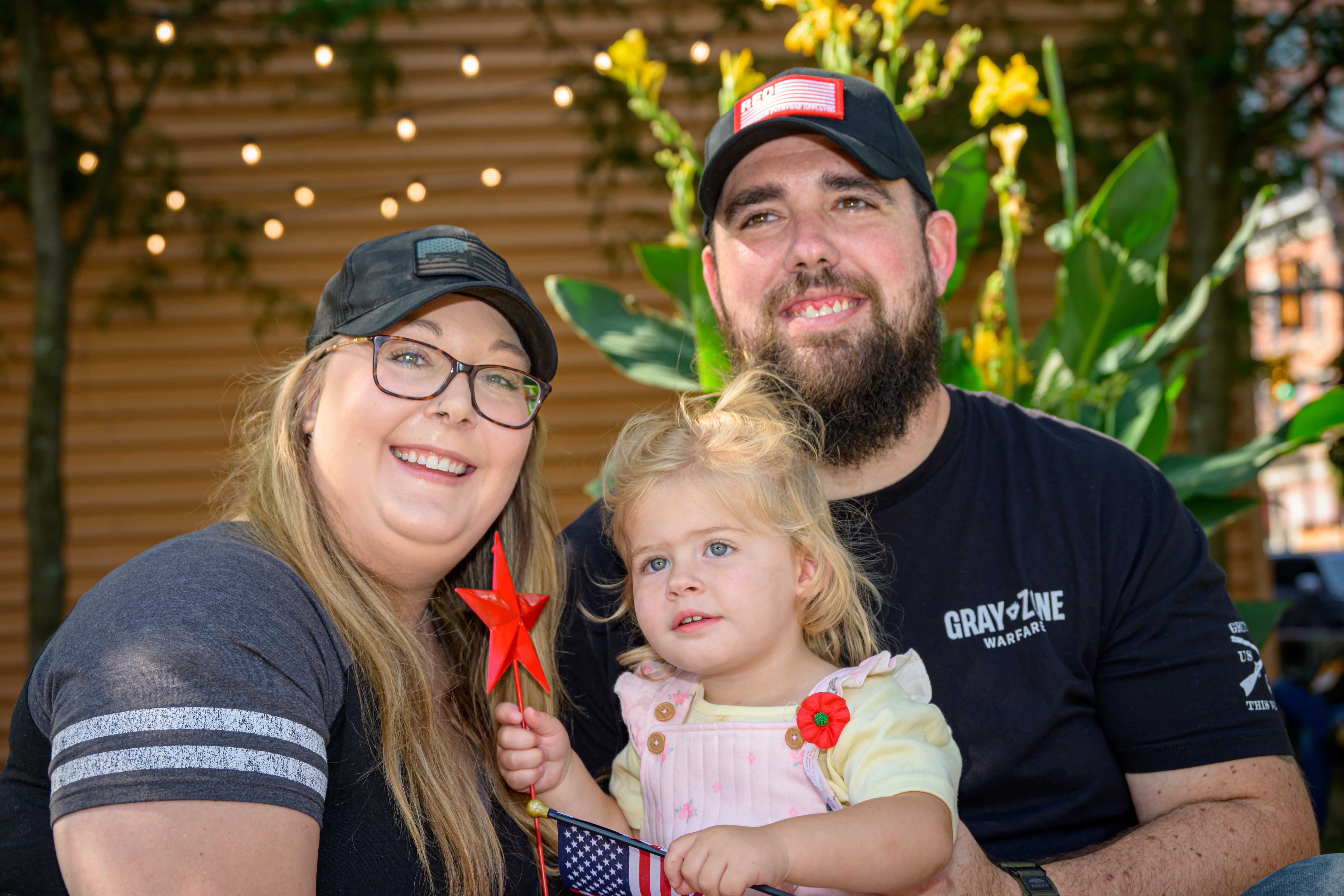 A veteran and his wife smile with their toddler daughter at an outdoor community event, the child holding a small American flag