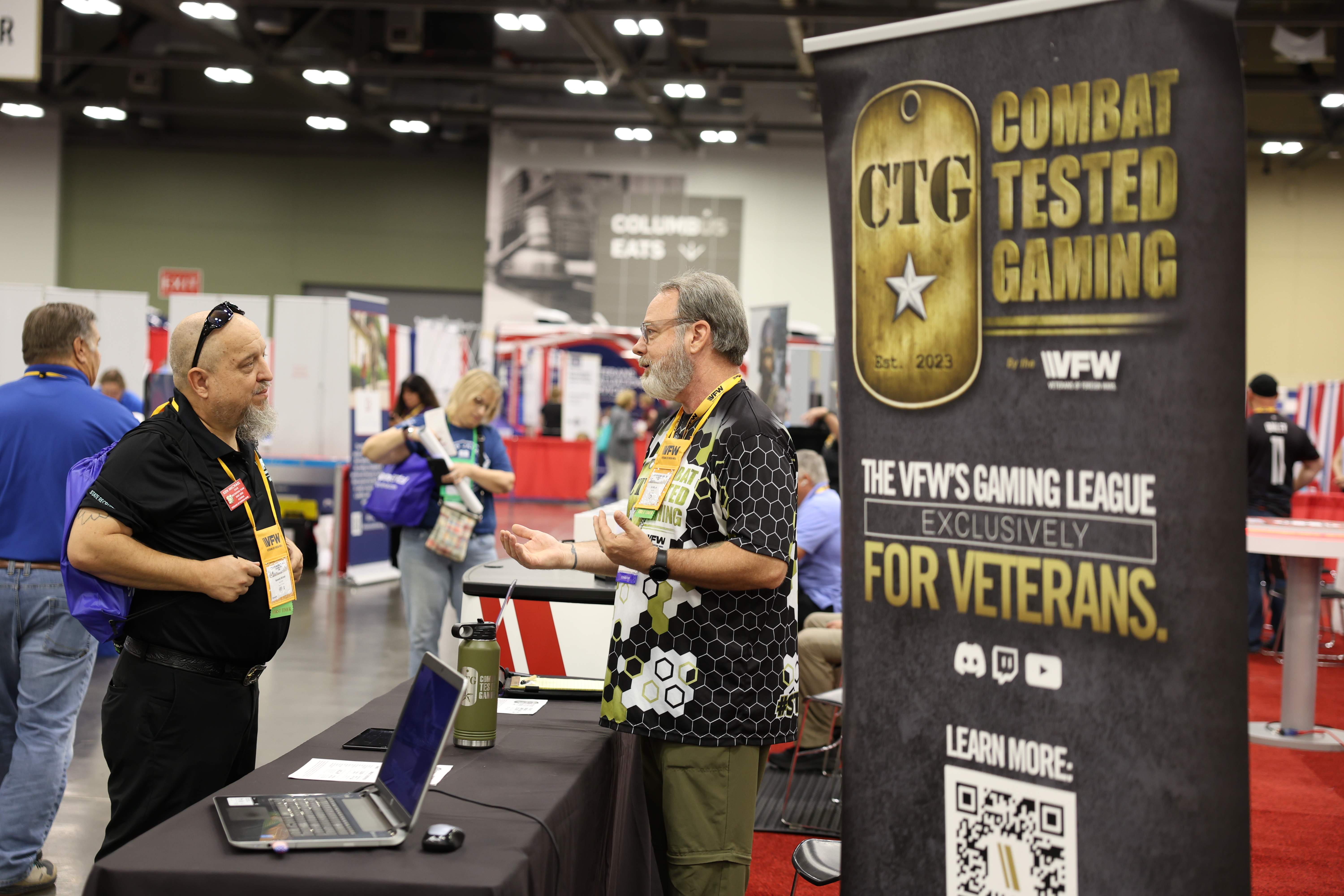 Two veterans in conversation at the Combat Tested Gaming booth beside a banner reading The VFW's gaming league exclusively for veterans
