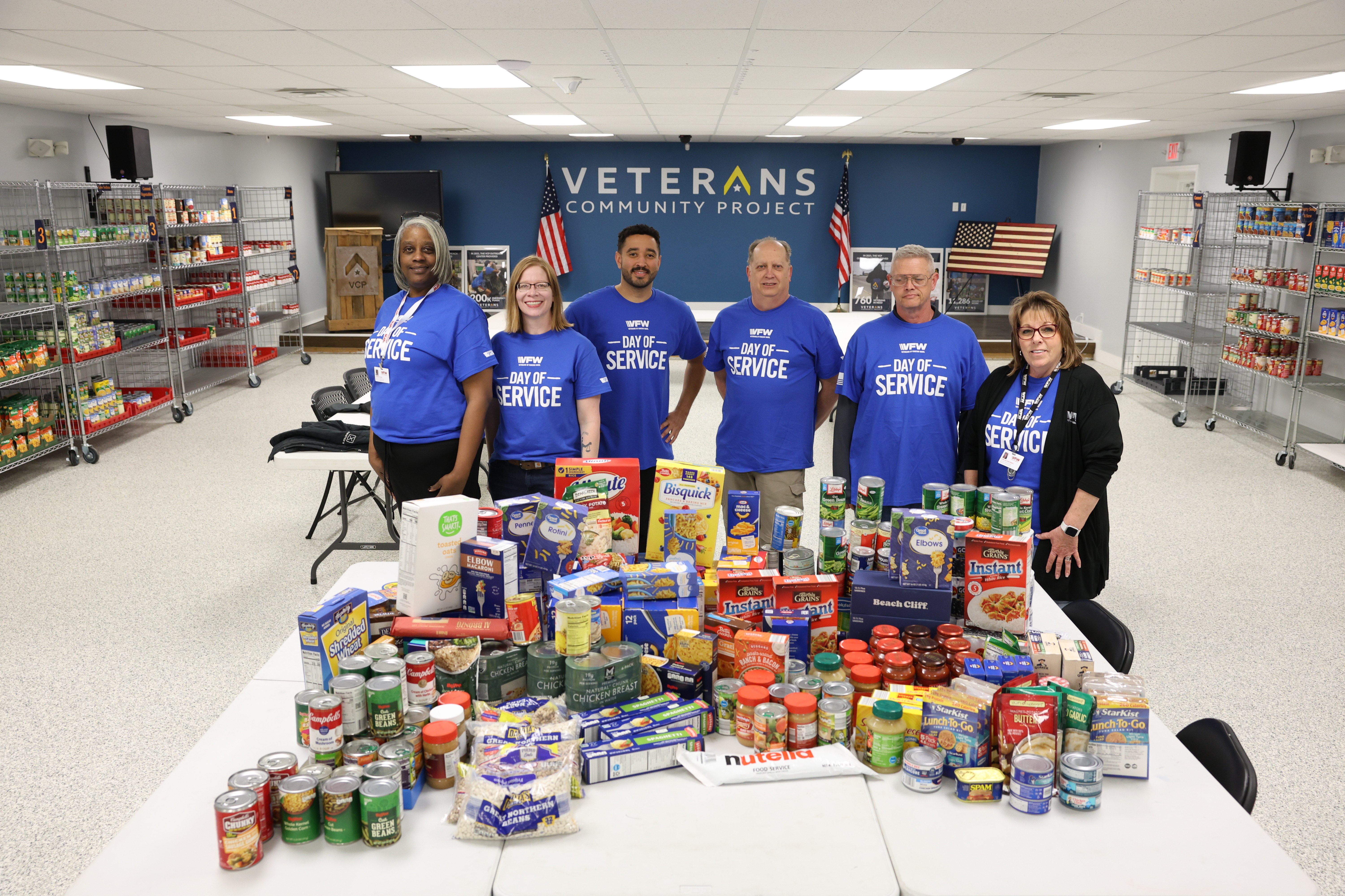 Six VFW volunteers in blue Day of Service shirts standing behind a table stacked with donated food at a Veterans Community Project pantry