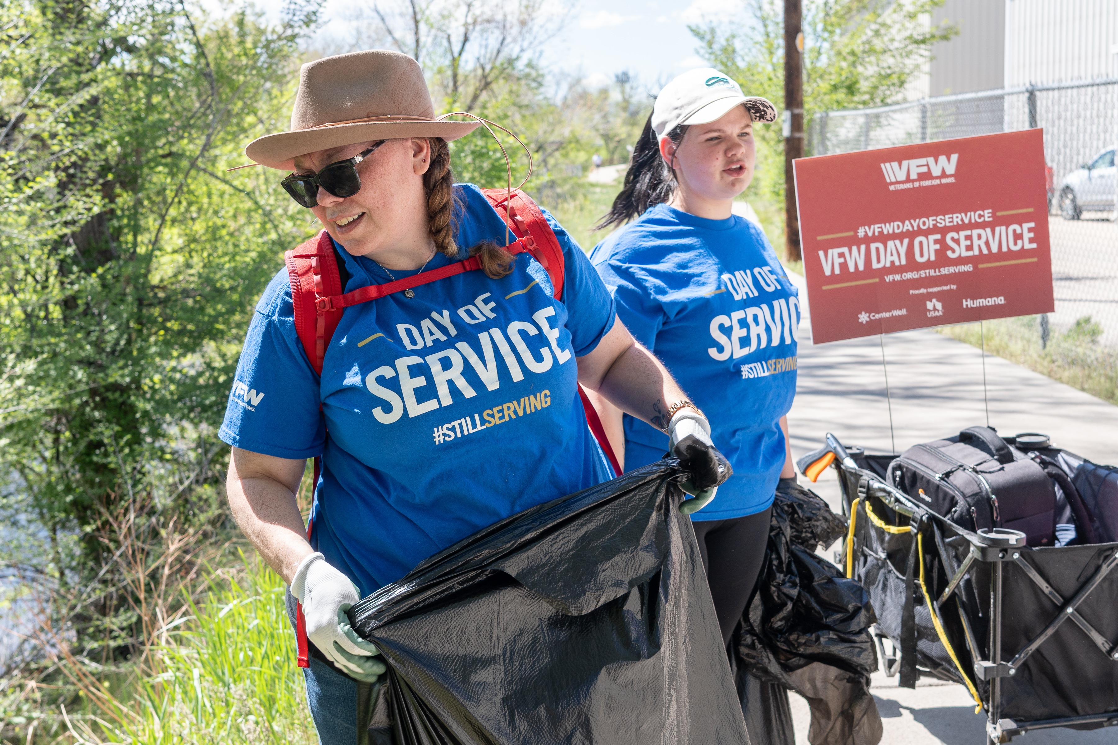 Two volunteers in blue VFW Day of Service shirts collecting litter into trash bags beside a VFW Day of Service sign.