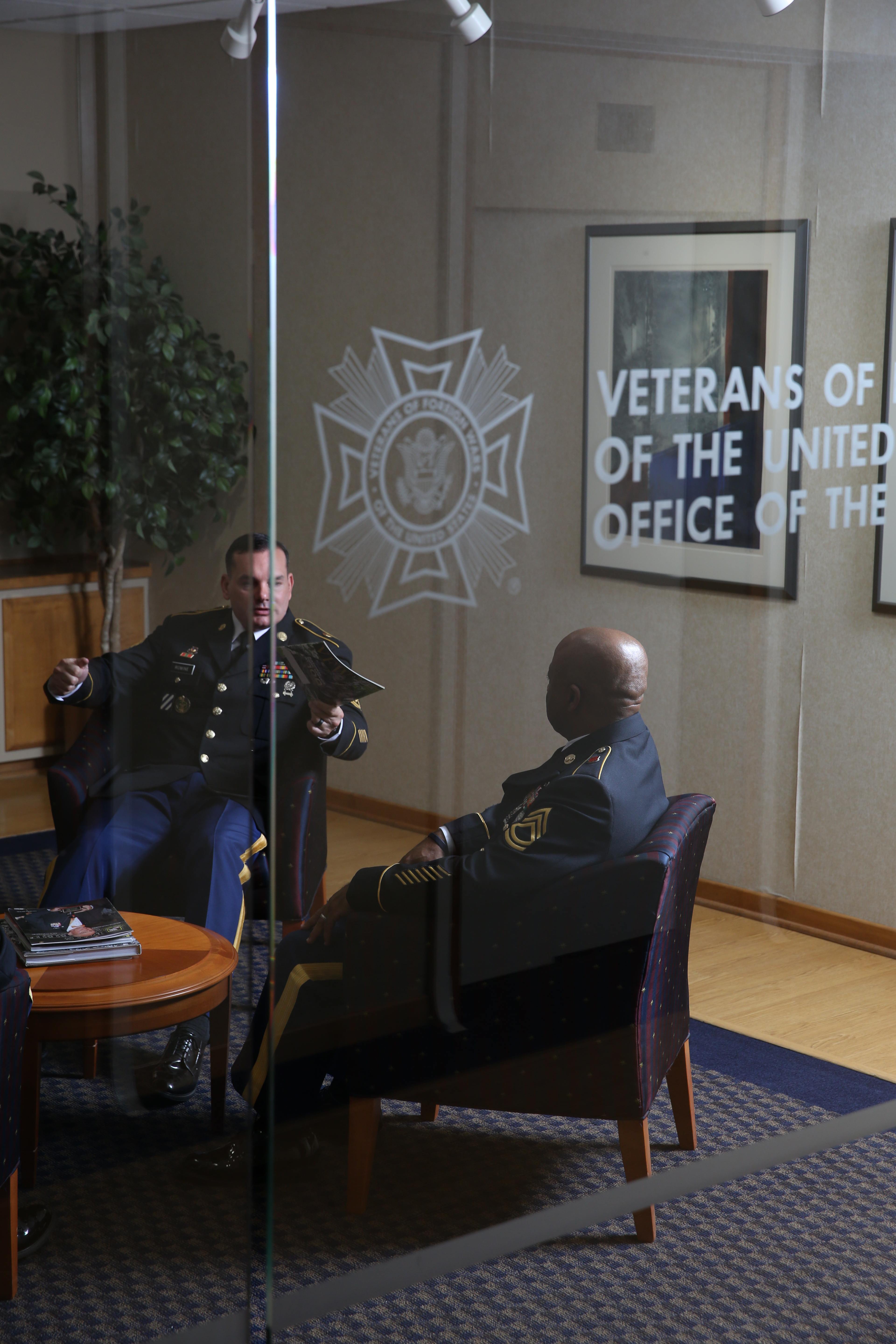 Two uniformed Army veterans seated in armchairs talking in a VFW office, viewed through glass etched with the VFW emblem